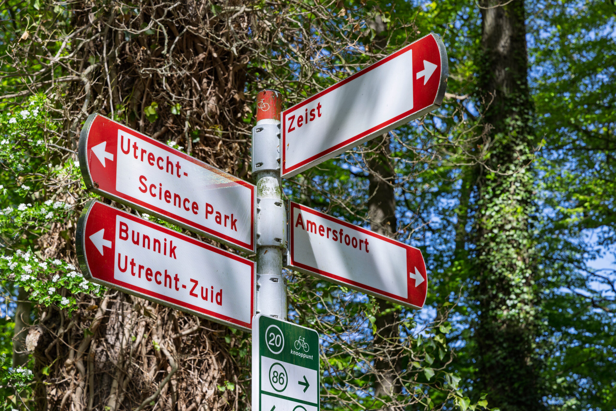 Zeist, Netherlands. Red and white cycling directional signs in a forest area showing routes to Utrecht Science Park, Amersfoort, Bunnik and Zeist city center.